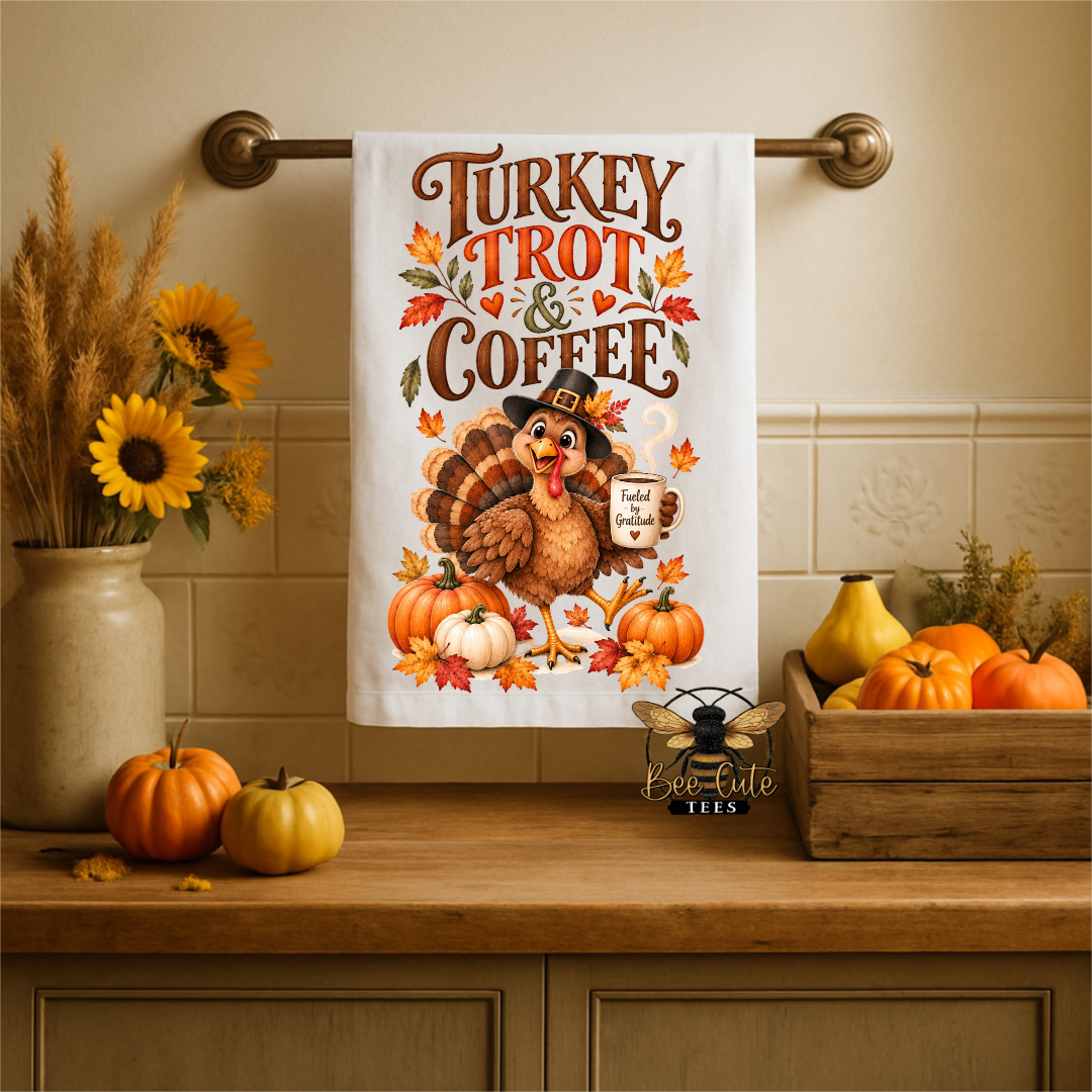 A white kitchen towel with a Thanksgiving-themed illustration hanging on a brass towel bar. The towel features the text 'Turkey Trot & Coffee' in brown lettering, surrounded by autumn leaves, pumpkins, and a turkey holding a mug.