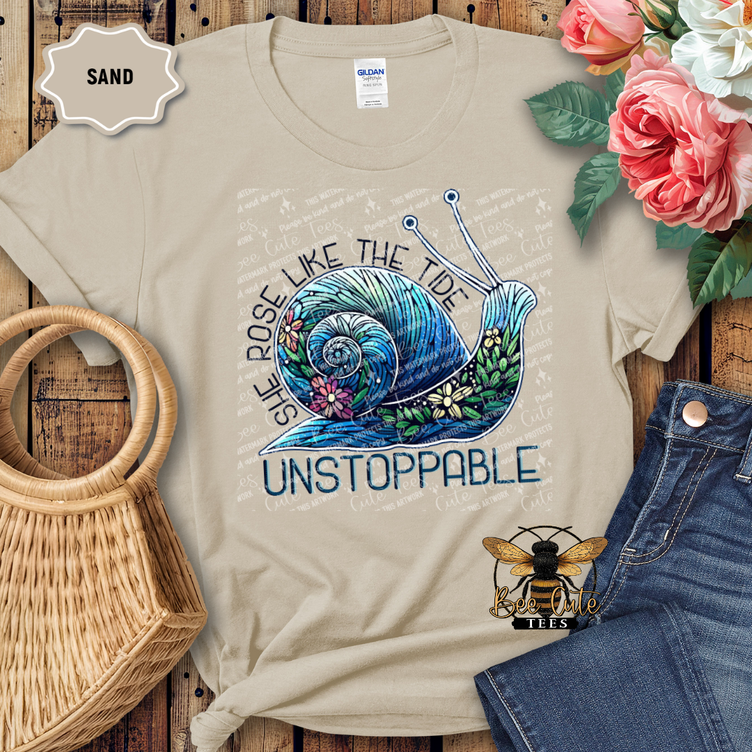 Beige t-shirt with a snail design and text on a wooden surface with flowers and a bag.