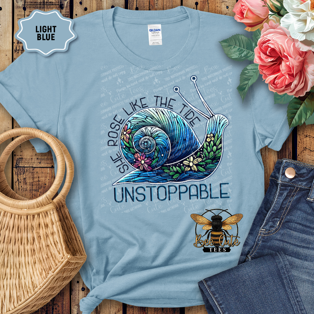 Light blue t-shirt with a graphic design featuring a wave and text, placed on a wooden surface with flowers and jeans.