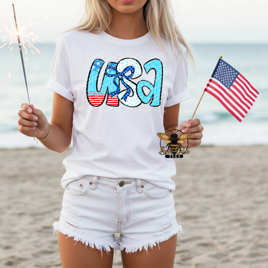 Person on a beach wearing a white t-shirt with 'USA' design, holding an American flag and a sparkler.