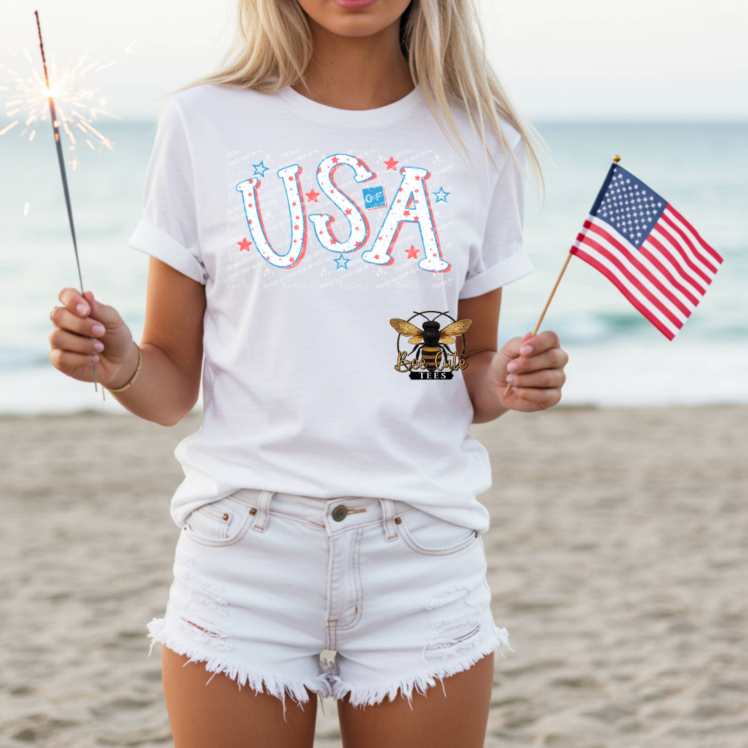 Person on a beach wearing a white t-shirt with 'USA' and a bee logo, holding an American flag and a sparkler.