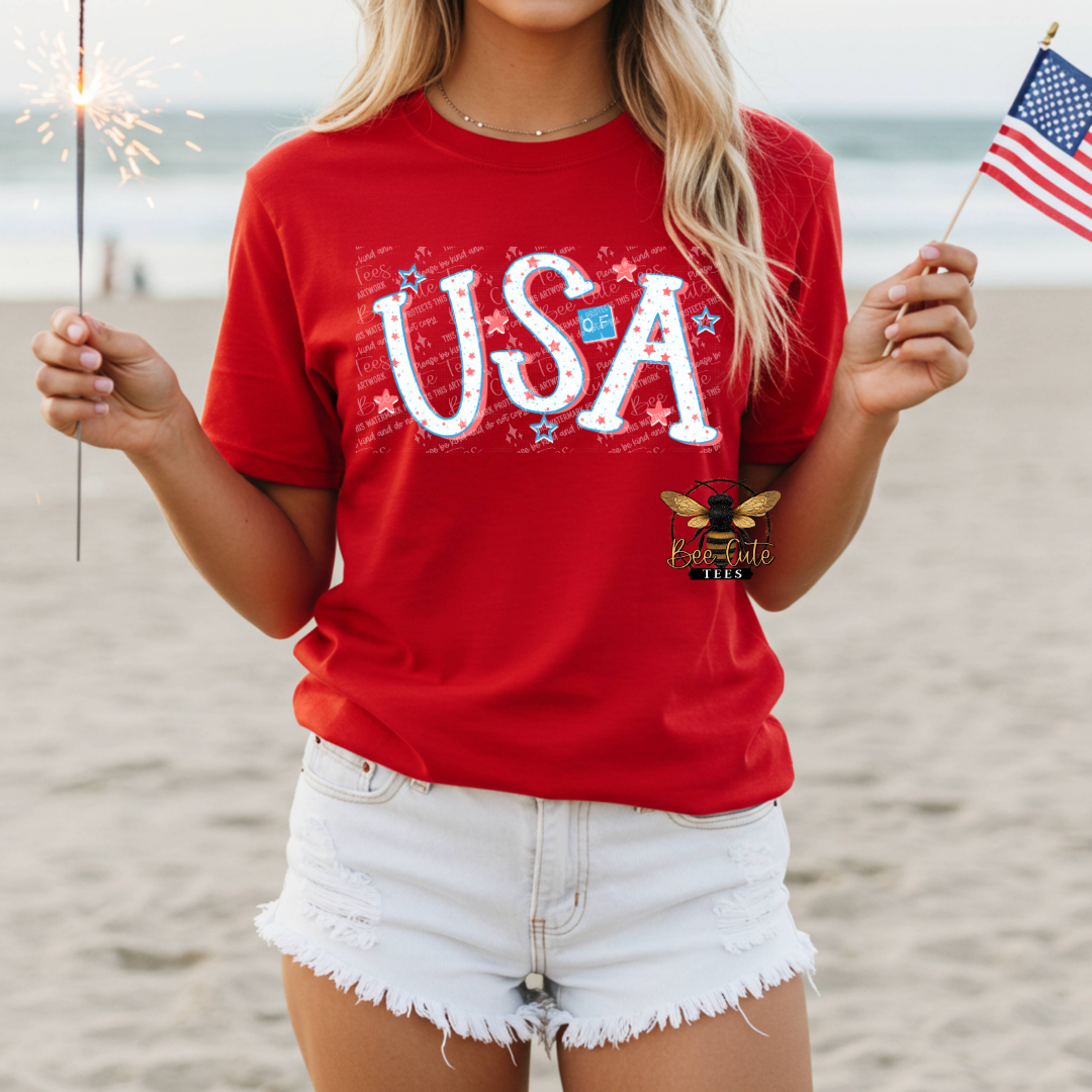 Person wearing a red 'USA' t-shirt on a beach with sparklers and an American flag.