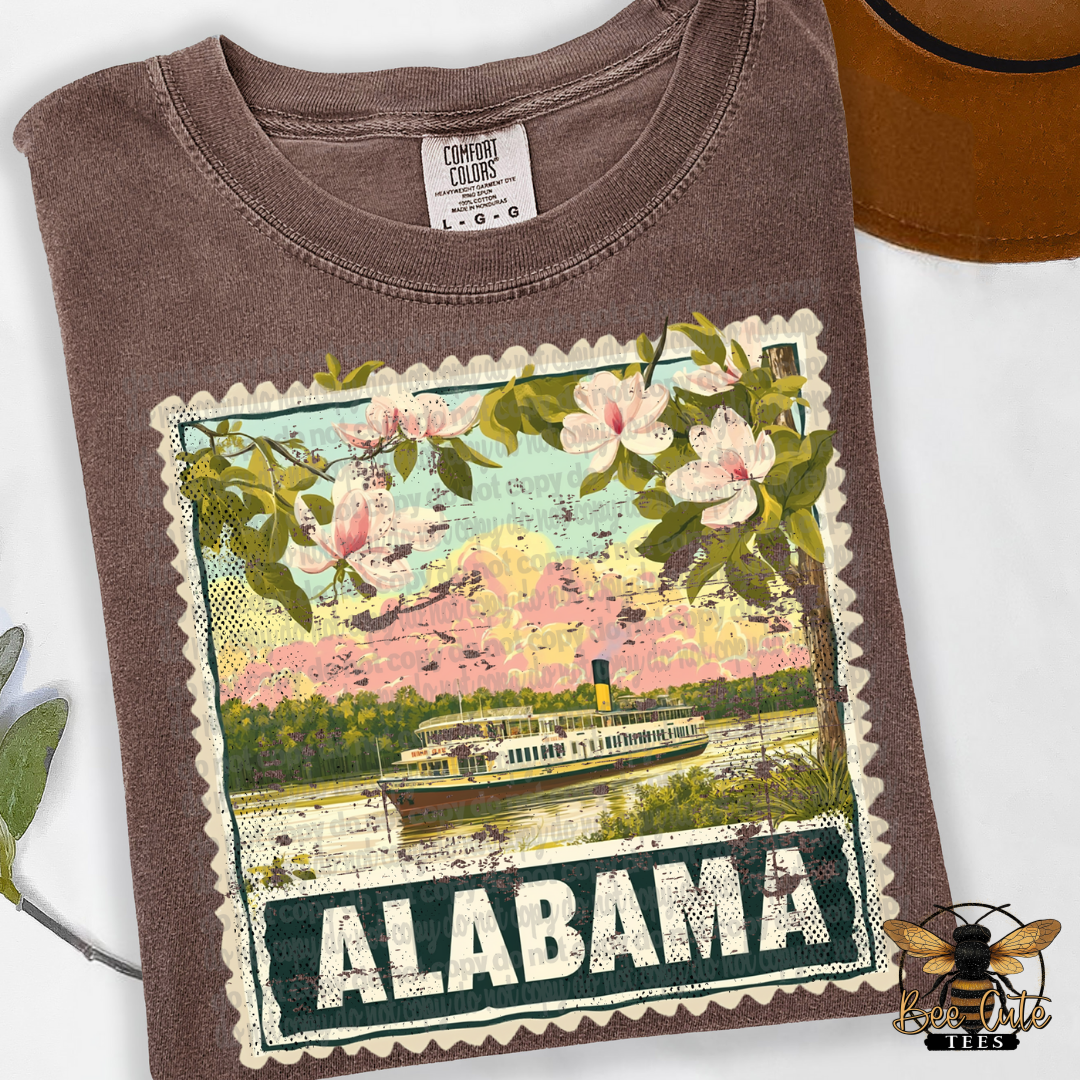 Brown t-shirt with a graphic design of a river scene and 'Alabama' text, placed on a white surface.