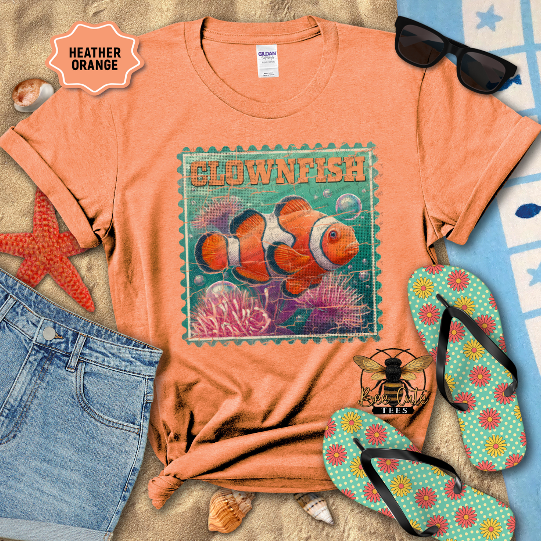 Orange t-shirt with clownfish design, sunglasses, jeans, starfish, and flip-flops on a sandy surface.