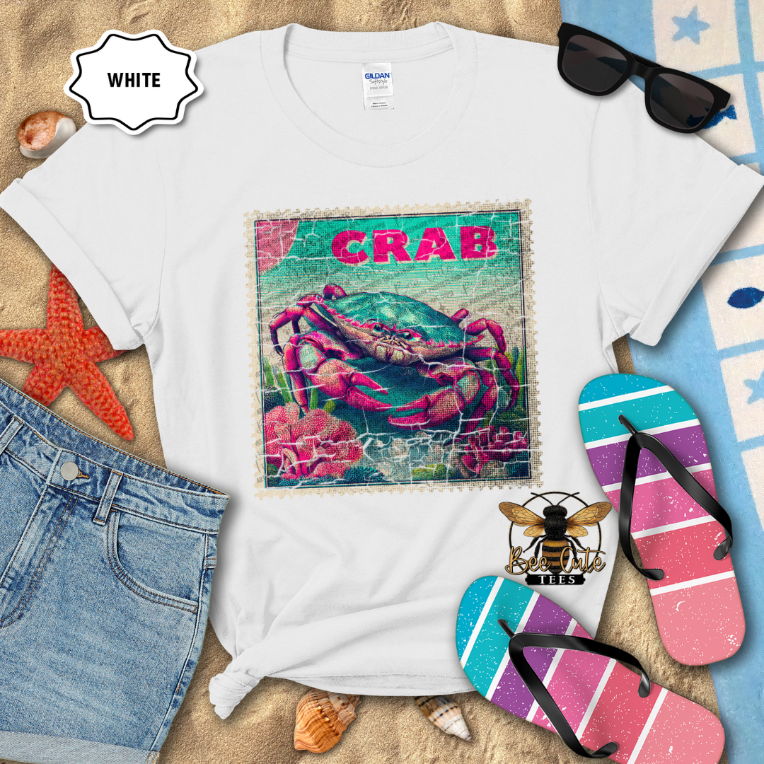 White t-shirt with a colorful crab design on a sandy surface with flip-flops and sunglasses.