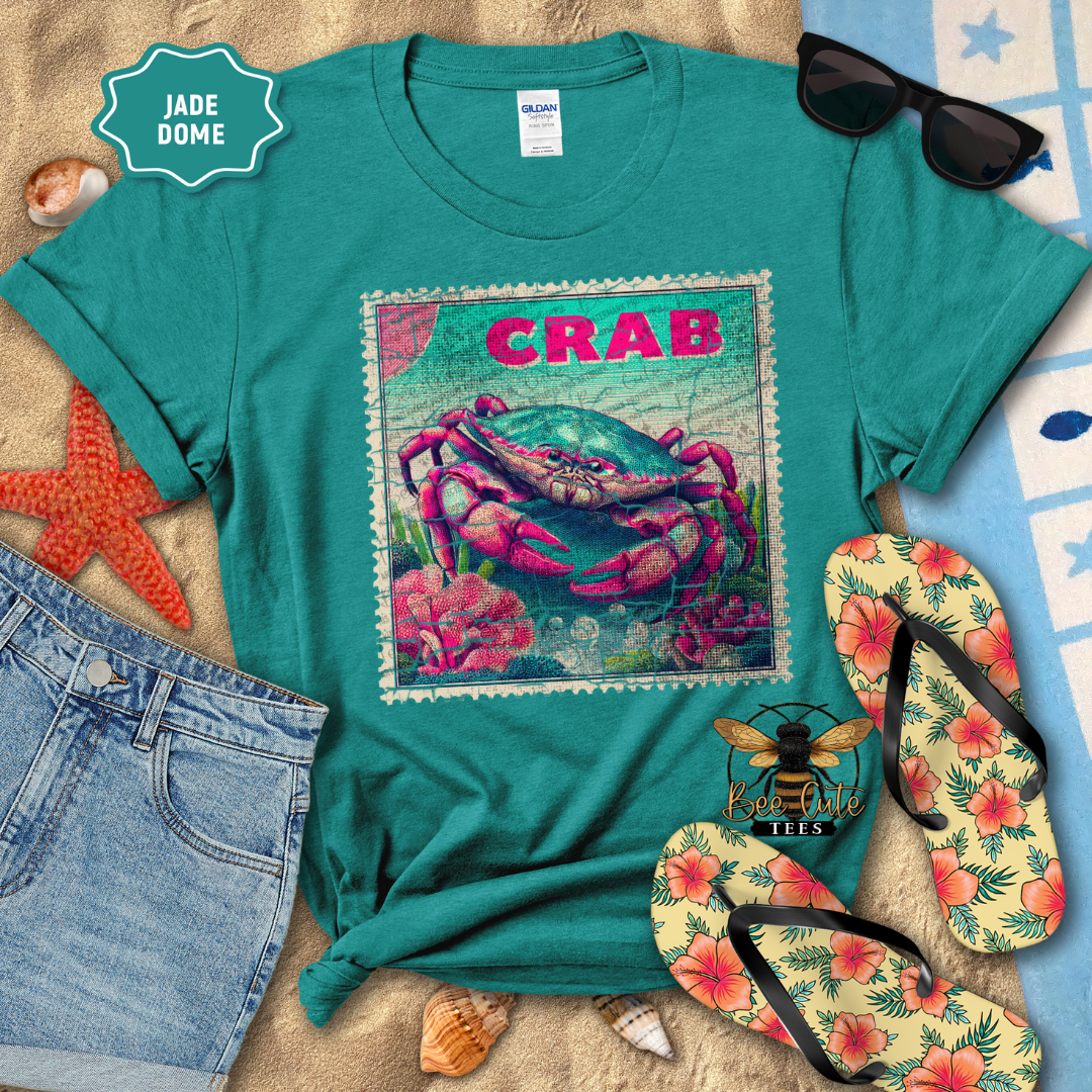 Teal t-shirt with a colorful crab design on a sandy surface with flip-flops and sunglasses.