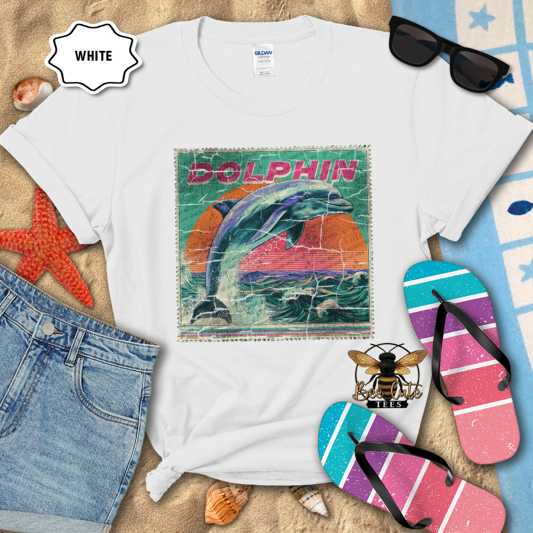 White t-shirt with a dolphin graphic design on a beach-themed background with sunglasses, flip-flops, and denim shorts.