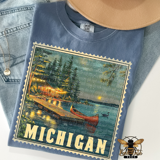 Blue t-shirt with a graphic of a lake scene and 'Michigan' text, displayed with jeans and a hat.