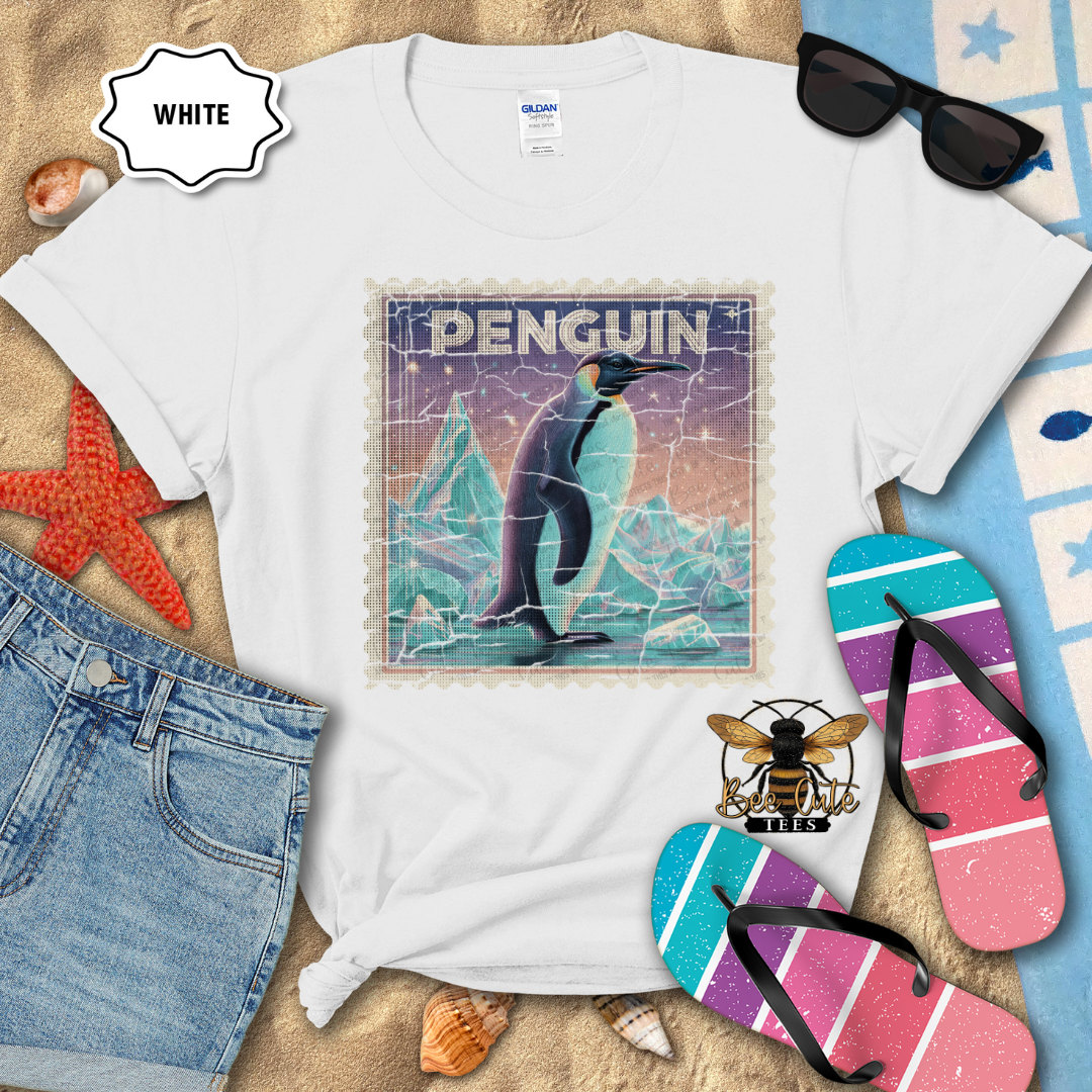 White t-shirt with penguin graphic design on a sandy surface with beach accessories.