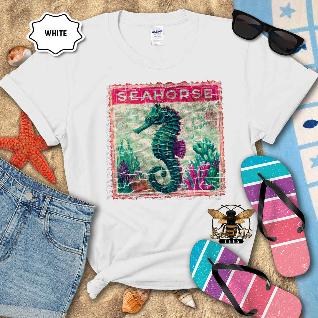 White t-shirt with seahorse design surrounded by beach-themed items on a sandy surface.