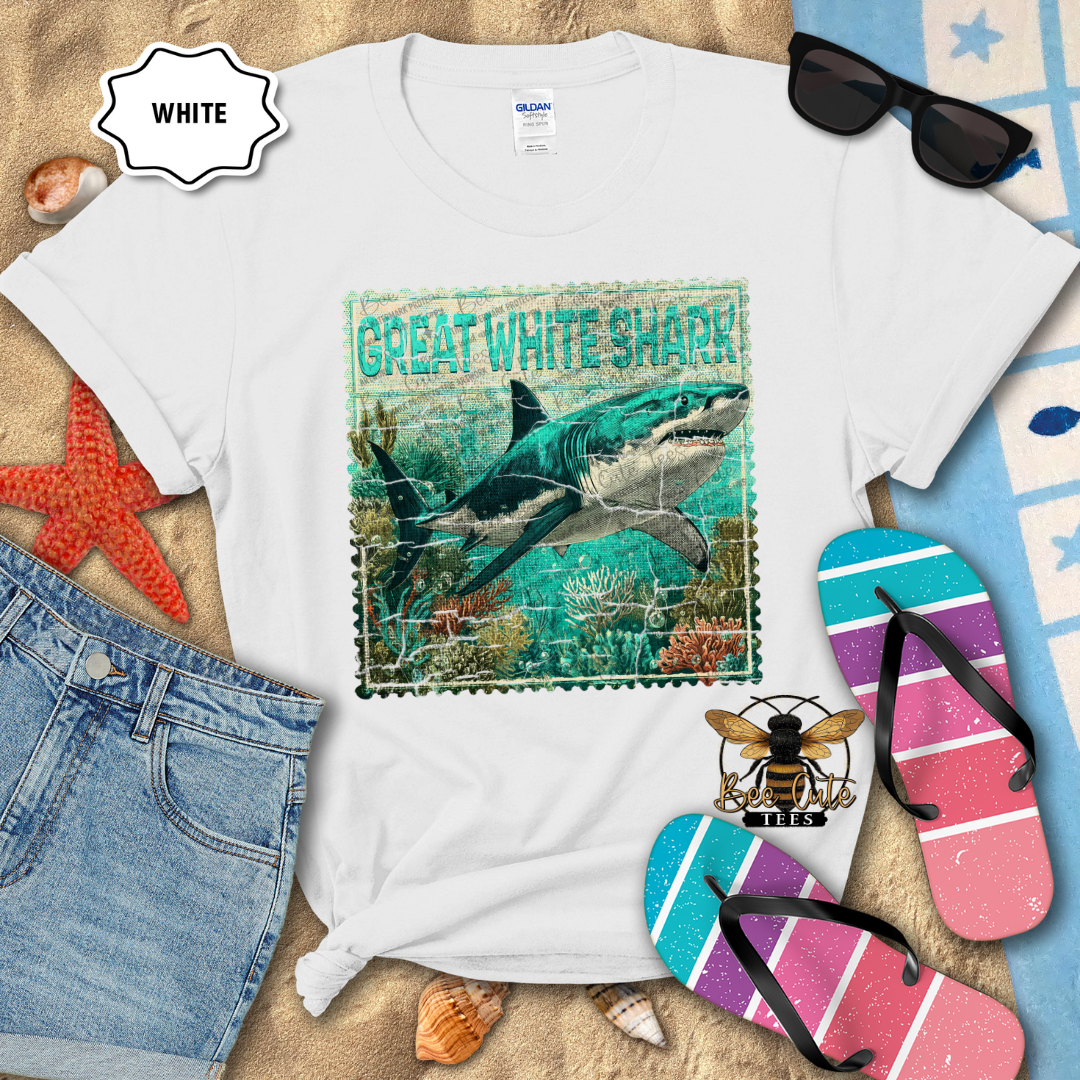 White t-shirt with a Great White Shark design on a sandy surface with flip-flops and sunglasses.