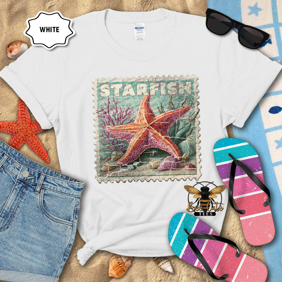 White t-shirt with starfish design on a sandy surface with beach accessories.