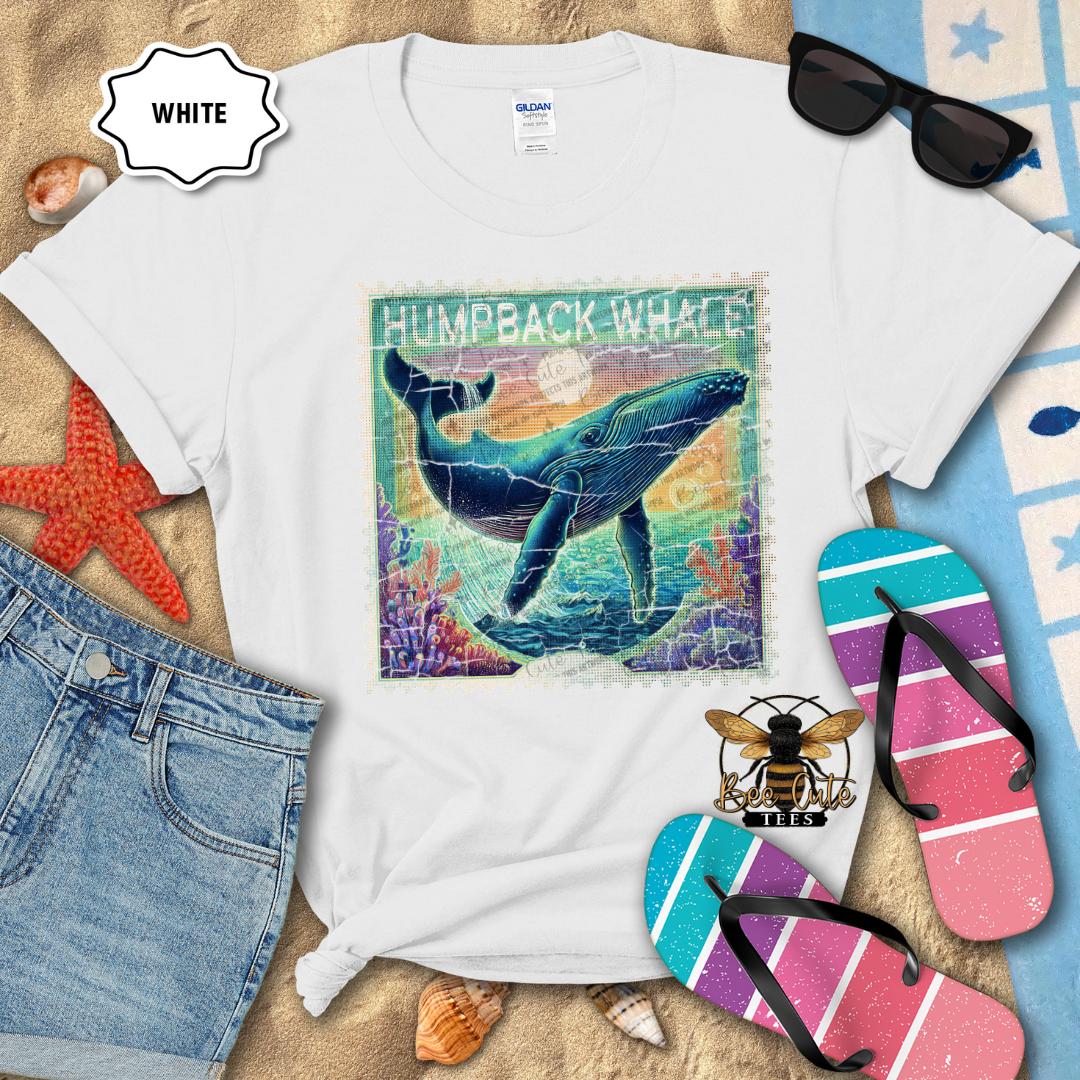 White t-shirt with humpback whale graphic on a sandy surface with beach accessories.