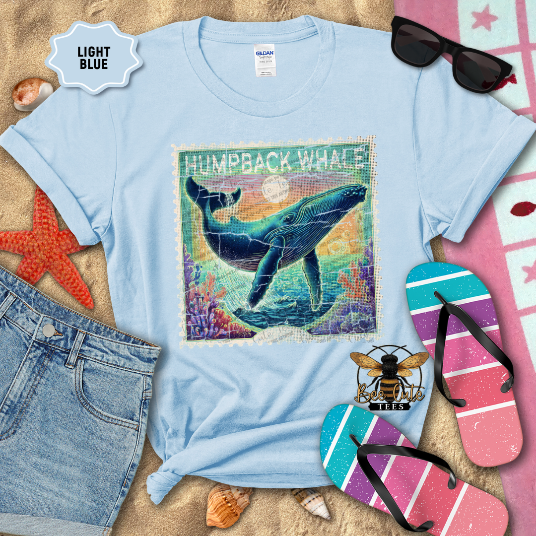Light blue t-shirt with humpback whale graphic on a sandy background with beach accessories.