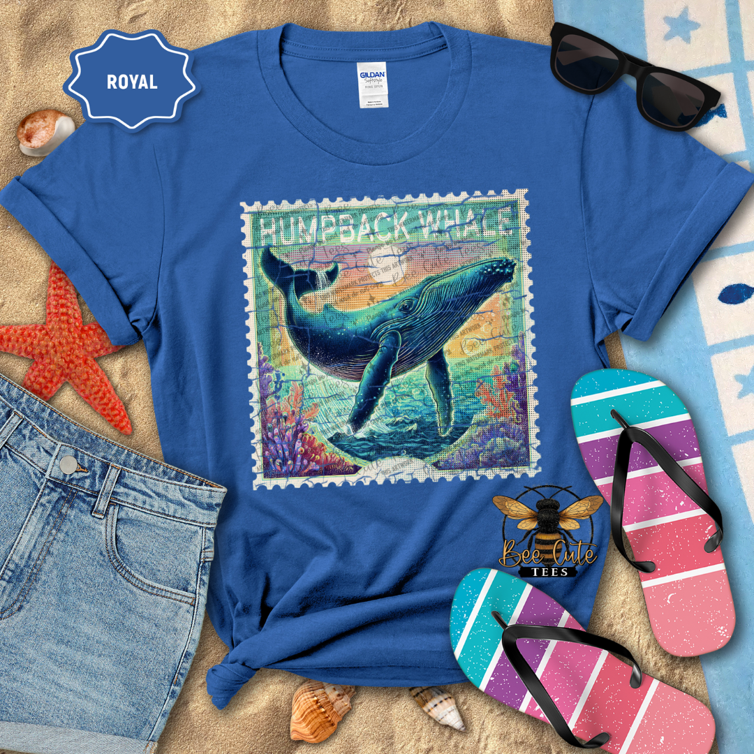 Blue t-shirt with humpback whale design on a sandy surface with beach accessories.