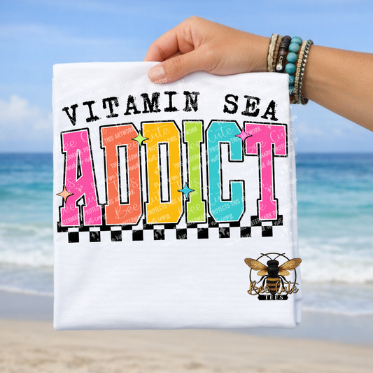 Hand holding a t-shirt with 'Vitamin Sea Addict' text against a beach background