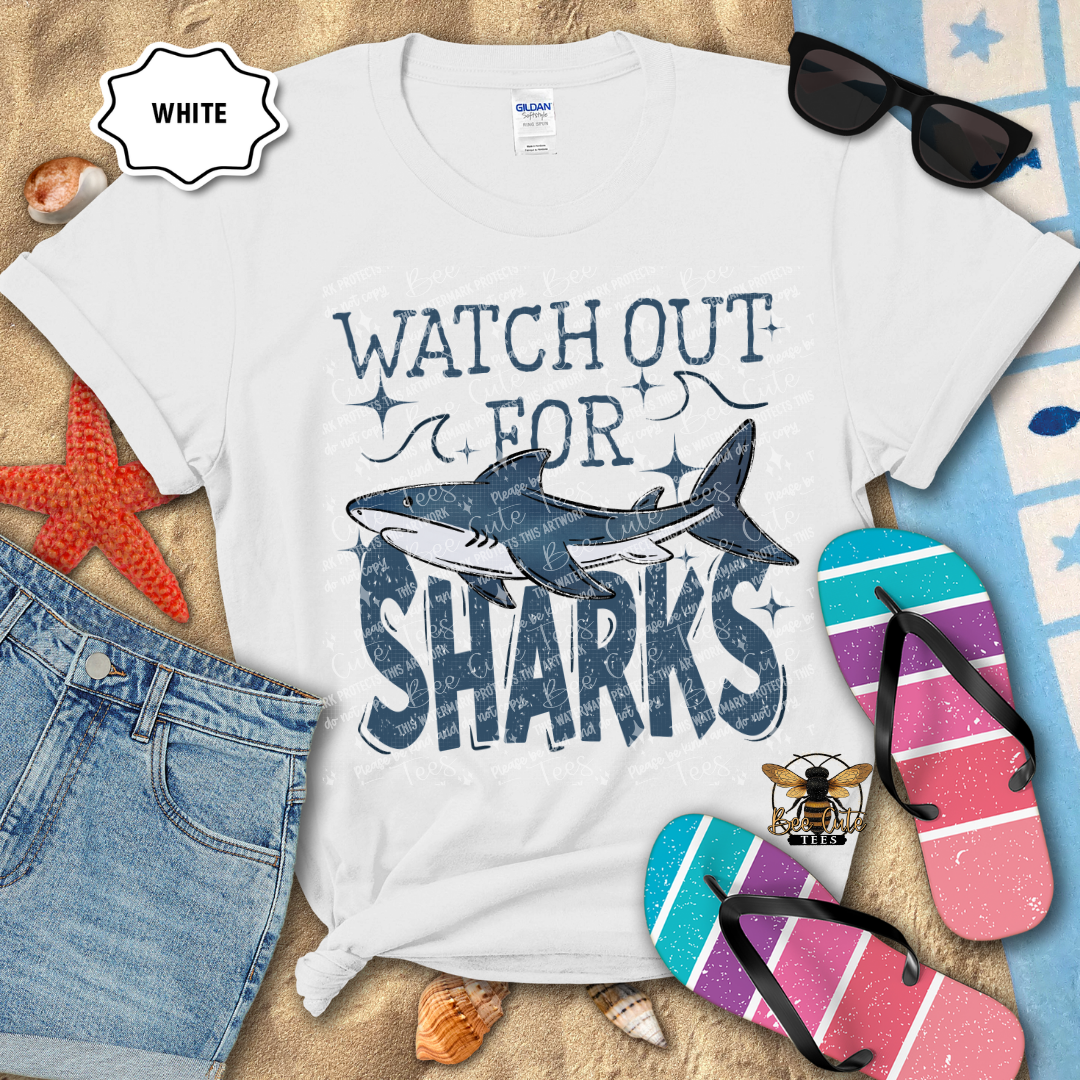 White t-shirt with 'Watch Out for Sharks' design on a beach-themed background with sunglasses, flip-flops, and starfish.