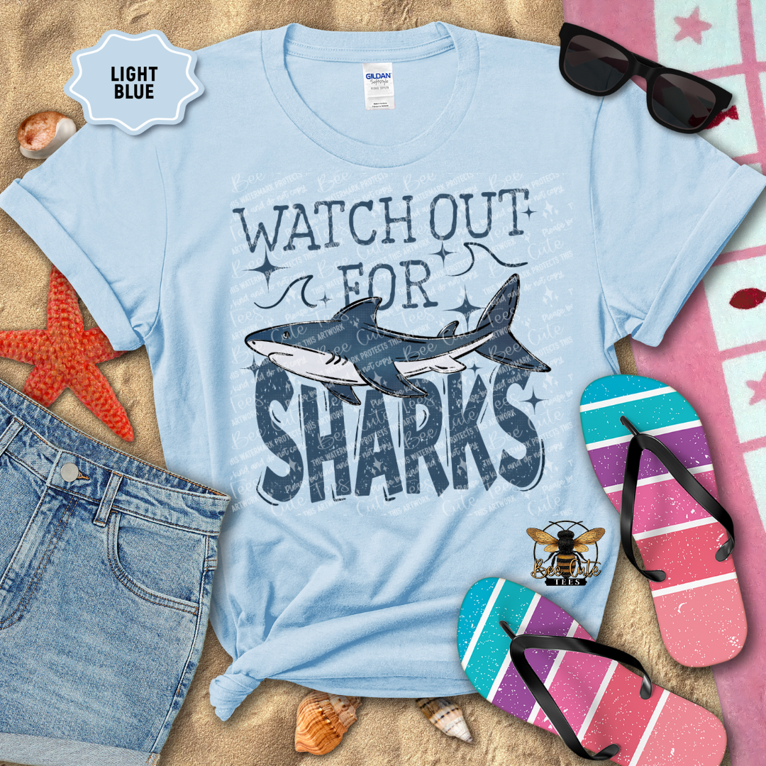Light blue t-shirt with 'Watch Out for Sharks' graphic on a beach-themed background with sunglasses, flip-flops, and starfish.