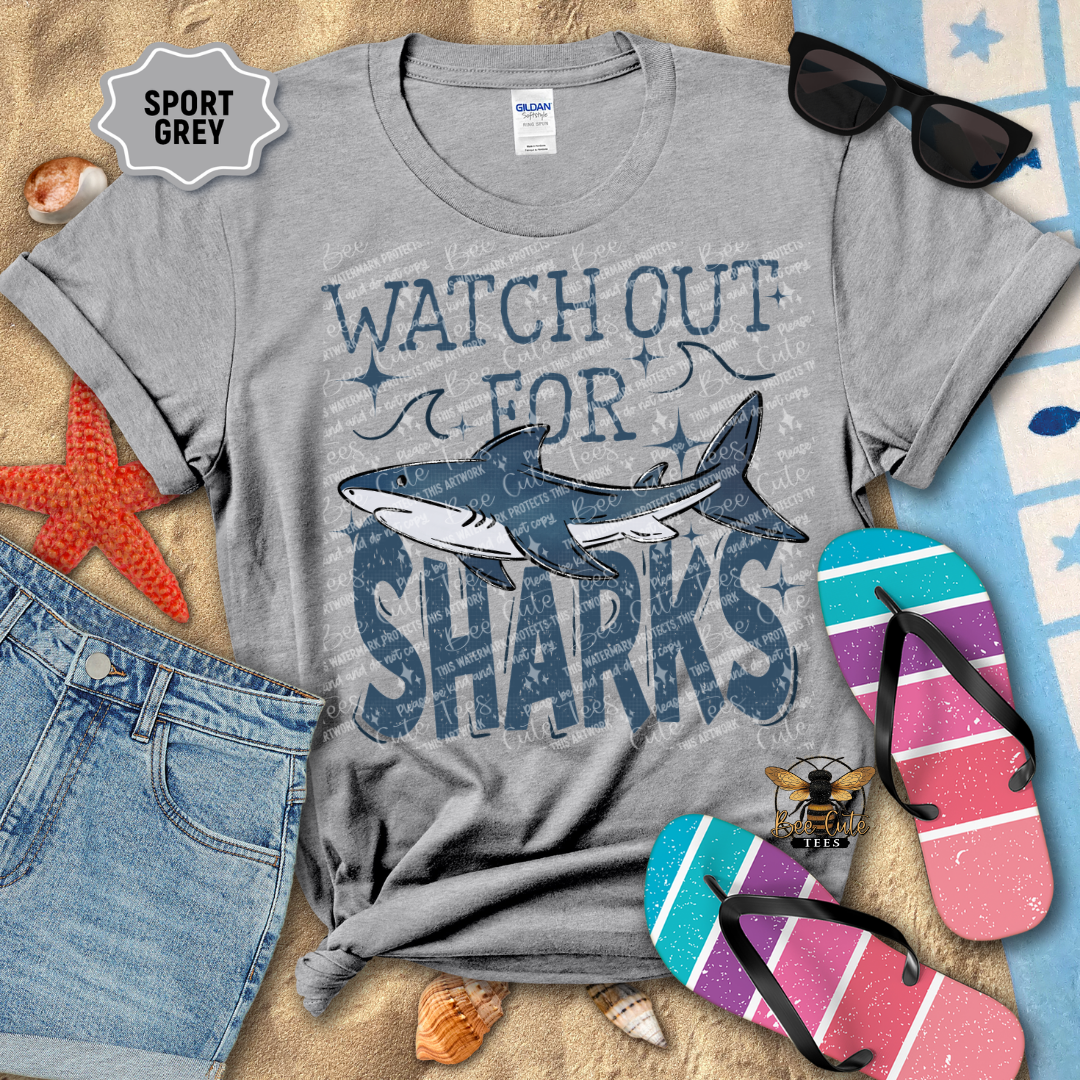 Gray t-shirt with shark graphic and text 'Watch Out for Sharks' on a beach-themed background.