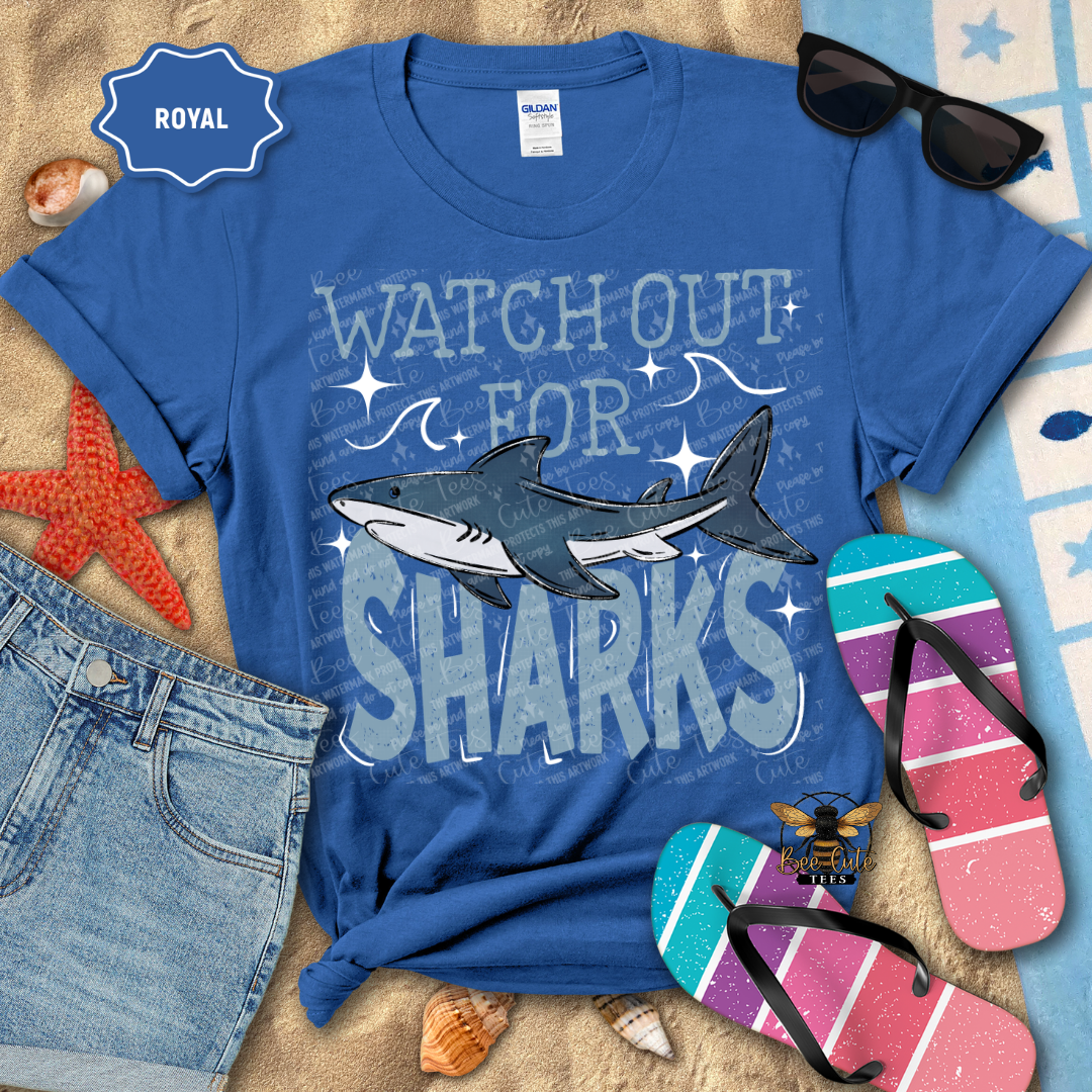 Blue t-shirt with shark design and text, sunglasses, flip-flops, and denim shorts on a beach-themed background.