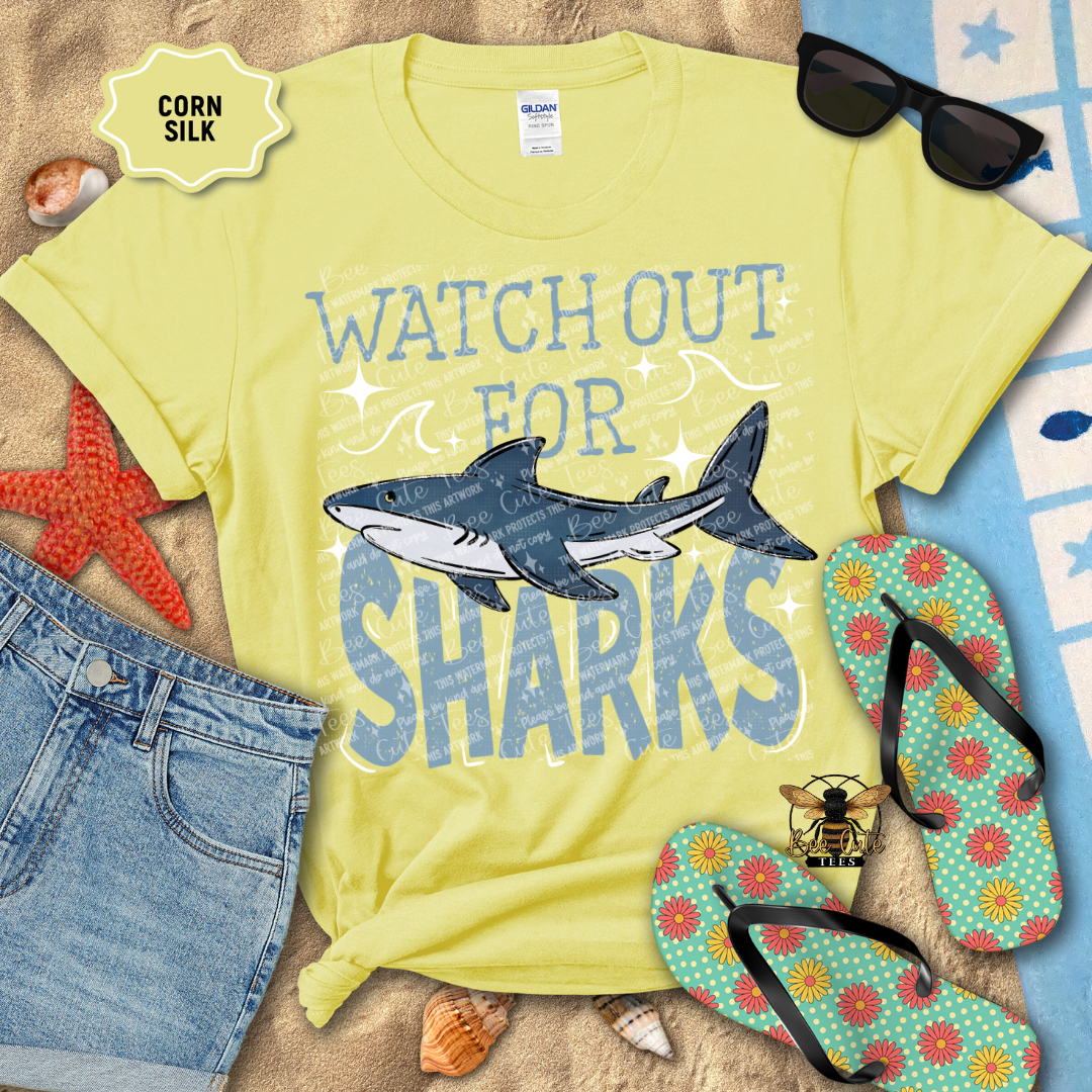 Yellow t-shirt with shark graphic and text 'Watch Out for Sharks' on a beach-themed background.