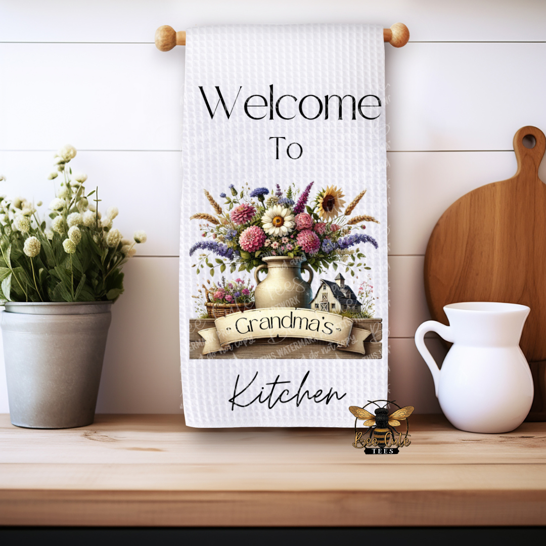 Kitchen towel with floral design and 'Welcome to Grandma's Kitchen' text on a wooden shelf.