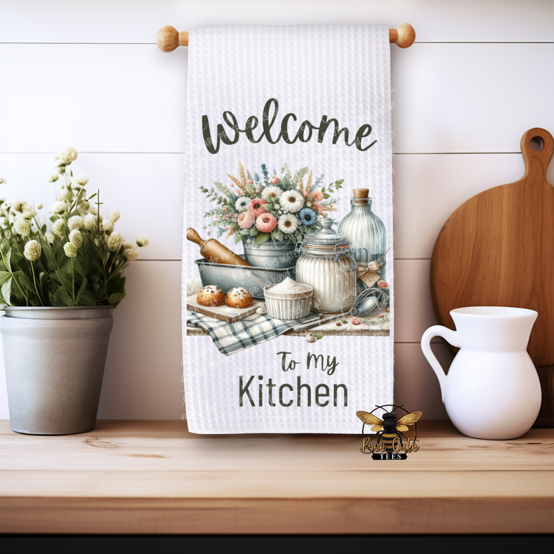 Kitchen towel with 'Welcome to my Kitchen' text and a rustic kitchen scene, hanging on a wooden rack.