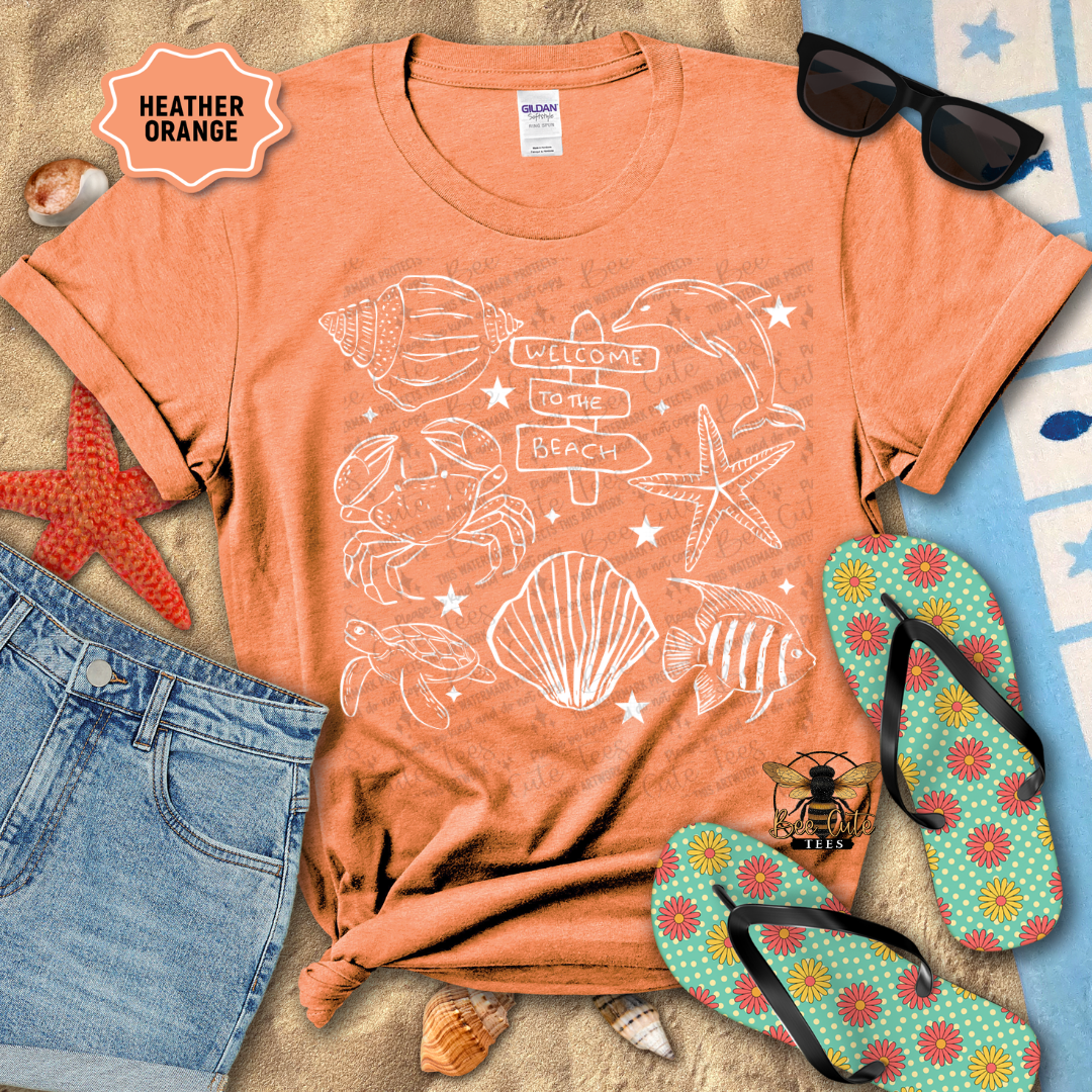 Orange t-shirt with beach-themed design, sunglasses, jeans, and flip-flops on a sandy surface.