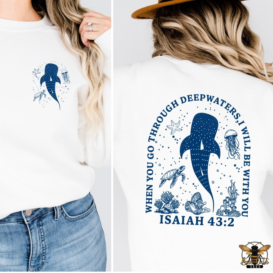 Split-view image of a white crewneck sweatshirt showcasing both the front and back designs. The front features a small blue whale shark surrounded by a sea turtle, jellyfish, starfish, and bubbles on the left chest. The back displays a larger whale shark with ocean life and the Bible verse, "When you go through deep waters, I will be with you – Isaiah 43:2," in bold blue lettering. The faith-inspired design is modeled with blue jeans and a tan wide-brim hat, creating a coastal, nautical look.
