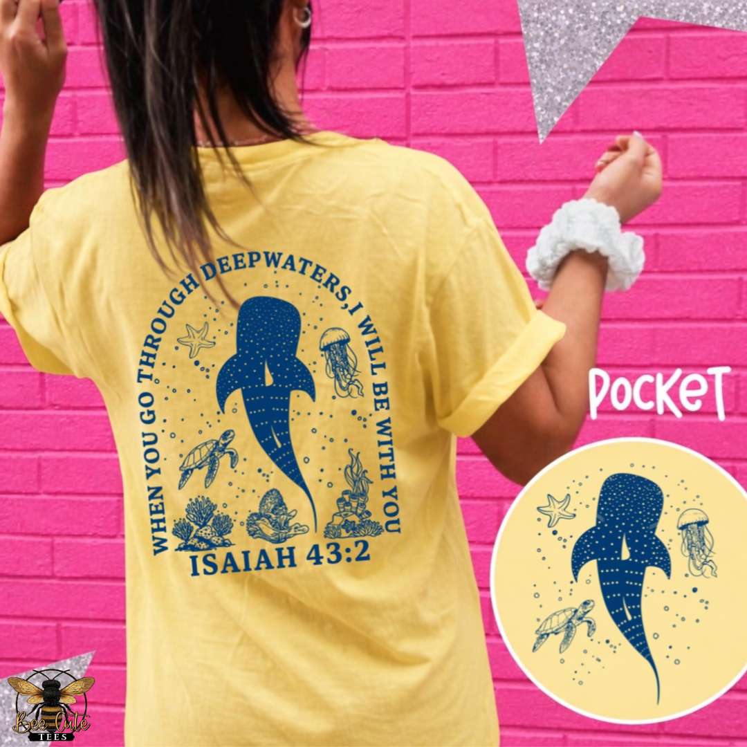 A person wearing a yellow T-shirt shown from the back against a bright pink brick wall. The shirt has a large navy blue whale graphic with ocean-themed illustrations and the text 'WHEN YOU GO THROUGH DEEPWATERS I WILL BE WITH YOU ISAIAH 43:2'.