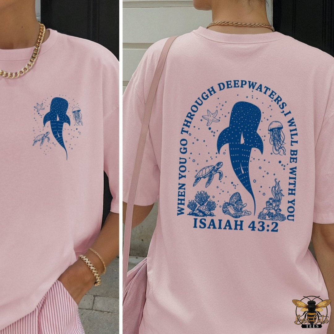A light pink graphic T-shirt shown on a model from both the front and back. The front features a blue whale print on the left chest, while the back displays a large blue whale design surrounded by ocean-themed illustrations and the text 'WHEN YOU GO THROUGH DEEPWATERS I WILL BE WITH YOU ISAIAH 43:2'.