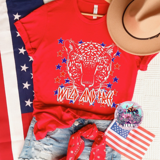 Red graphic t-shirt featuring a leopard face with stars and “Wild and Free” text, styled with distressed denim shorts, a red bandana, a tan hat, and American flag accents for a bold patriotic look.
