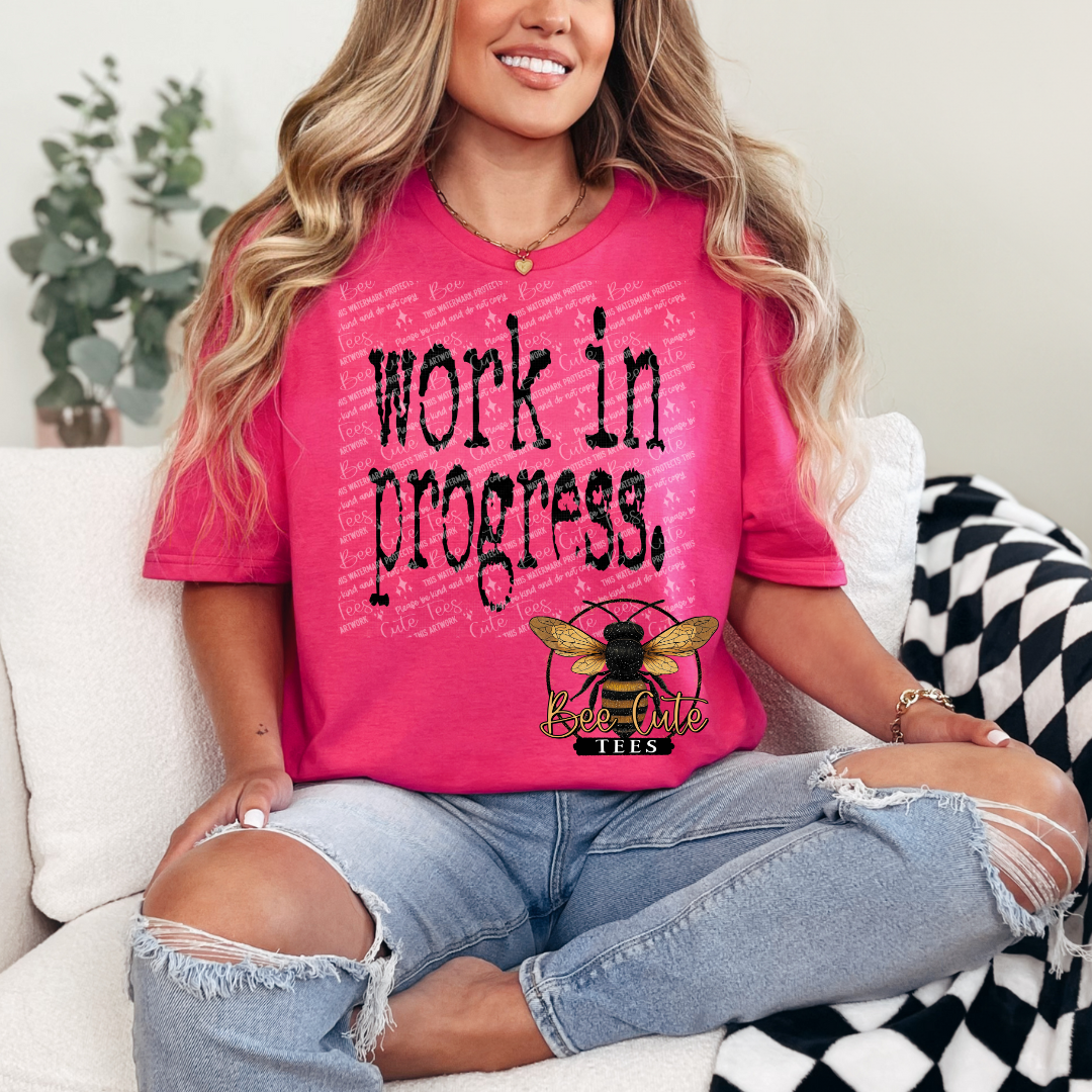 Person wearing a pink t-shirt with 'work in progress' text and a bee logo, sitting on a couch.