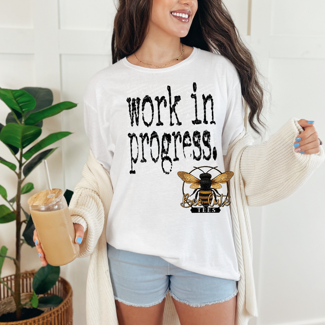 Woman wearing a white t-shirt with 'work in progress' text and a bee logo, holding a drink.