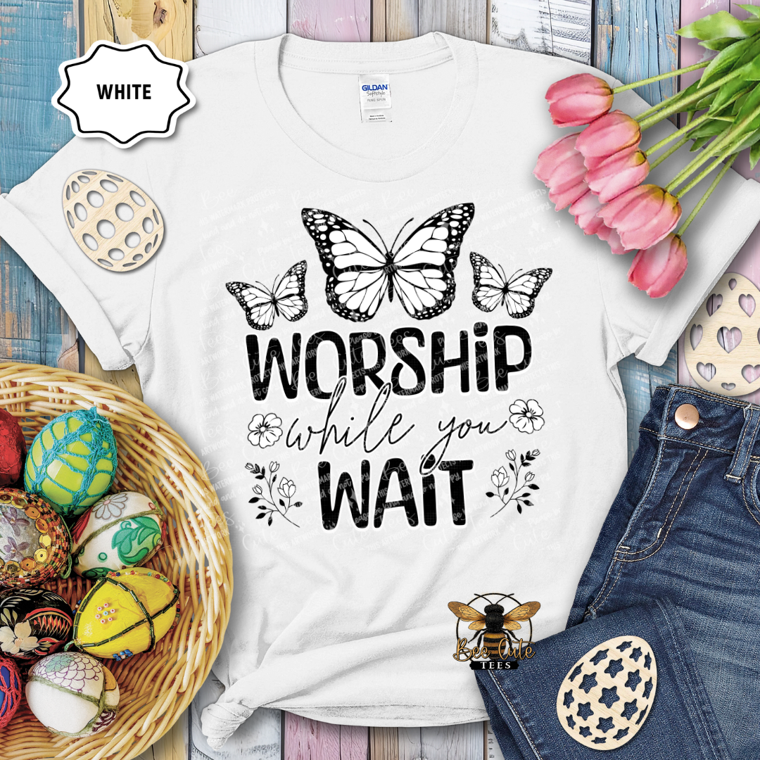 White t-shirt with 'Worship while you wait' text and butterfly design, surrounded by Easter decorations.