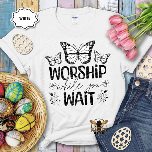White t-shirt with 'Worship while you wait' text and butterfly design, surrounded by Easter decorations.