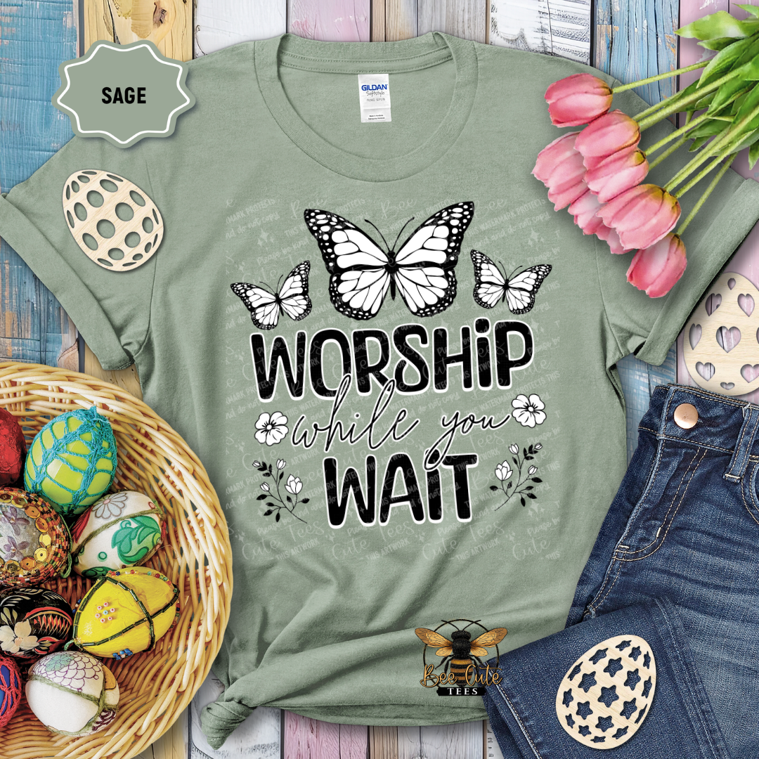 Green t-shirt with butterfly design and text 'Worship while you wait' on a wooden surface with Easter decorations.