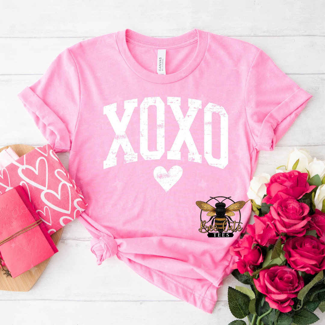 Pink t-shirt with 'XOXO' design on a white surface with flowers and a gift box.