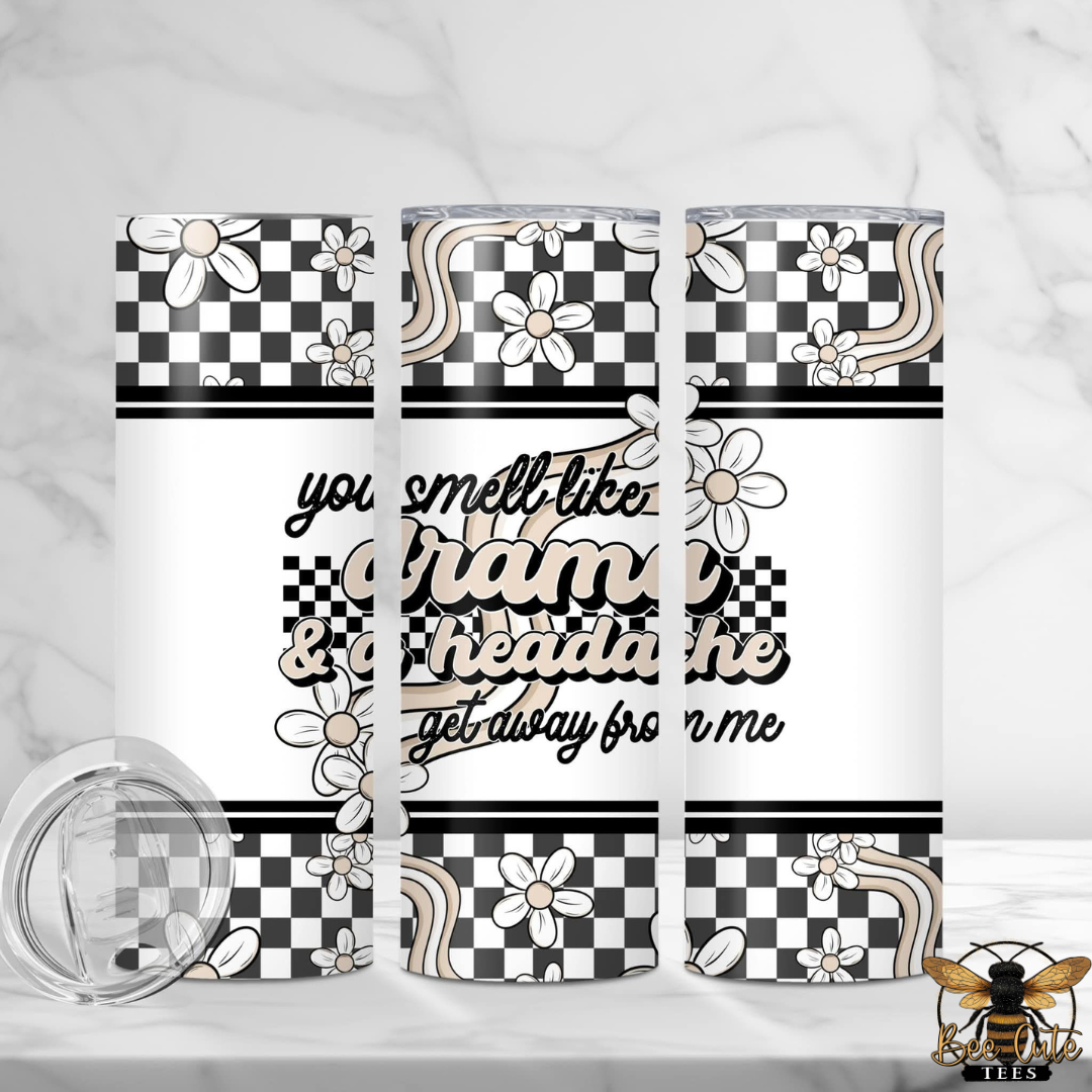 20 oz stainless steel insulated tumbler featuring a black, white, and beige retro-inspired wrap design with checkered patterns, daisies, and wavy accents. The center reads, "You smell like drama & a headache, get away from me" in layered black and beige lettering. The tumbler is displayed from multiple angles to show the full wrap design, with a clear sliding lid shown beside it.
