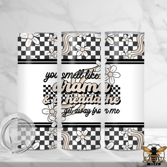 20 oz stainless steel insulated tumbler featuring a black, white, and beige retro-inspired wrap design with checkered patterns, daisies, and wavy accents. The center reads, "You smell like drama & a headache, get away from me" in layered black and beige lettering. The tumbler is displayed from multiple angles to show the full wrap design, with a clear sliding lid shown beside it.

